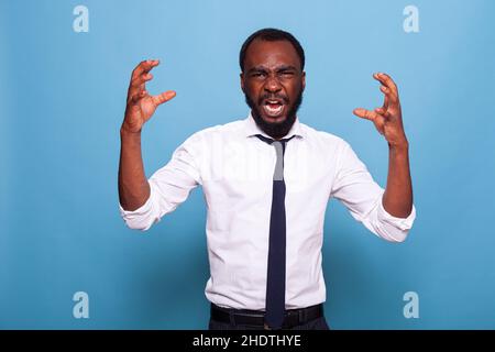 Porträt eines verärgerten afroamerikanischen Geschäftsmannes, der auf blauem Hintergrund schreit und die Hände hebt. Büroangestellter Buchhalter mit einem Nervenzusammenbruch. Frustrierter Mann mit aggressiver Reaktion. Stockfoto