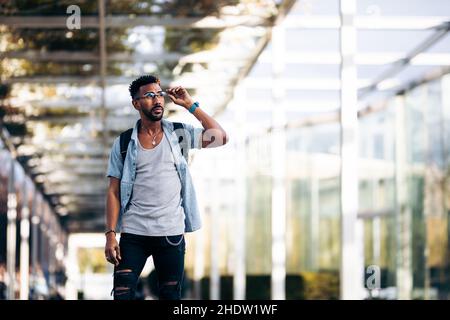 Schwarzer Mann mit Brille und Rucksack, der die Straße entlang läuft. Seine Brille mit der Hand aufheben. Stockfoto