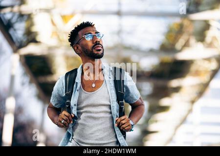 Schwarzer Mann mit Brille und Rucksack, der die Straße entlang läuft. Nehmen Sie die Griffe des Rucksacks mit den Händen. Stockfoto