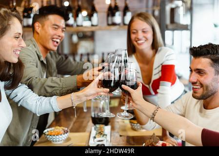 Eine bunte Gruppe multikultureller Freunde feiert im Restaurant gemeinsam lachend und scherzend, während sie sich mit einem Glas Rotwein anstoßen Stockfoto