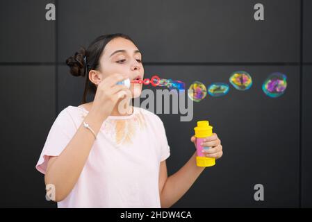 Teen Mädchen mit Haarbrötchen Blasen bunte Seifenblasen während der Sommerzeit gegen graue Wand genießen Stockfoto