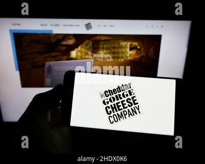 Person, die ein Mobiltelefon mit dem Logo der Cheddar Gorge Cheese Company Limited auf dem Bildschirm vor der Business-Webseite hält. Konzentrieren Sie sich auf die Telefonanzeige. Stockfoto