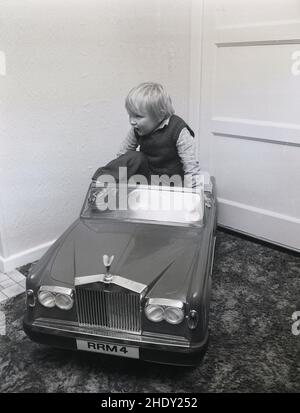 1980s, historisch, in einem Raum, klettert ein kleiner Junge in den Sitz seines Aufsitzwagens, eines Rolls Royce Corniche mit personalisierter Nummerierung RRM 4, England, Großbritannien. Dieser klassische Kinderpedalwagen wurde von Triang, einem führenden britischen Spielzeughersteller des Tages, hergestellt. Es hatte einen geformten Kunststoffkörper auf einem Stahlrahmen und einen Elektromotor. Als kleiner Junge ließ Prinz William ihm eines von Harrod's, dem berühmten Laden in Londons Knightsbridge, der sie ursprünglich verkaufte, geben. Stockfoto
