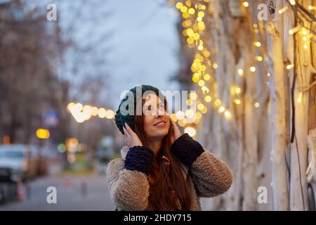 Verträumt schöne Frau lächelt in der Abendstraße. Lichter der Stadt. Winter-Freizeitkleidung. Festliche Stadt Stockfoto