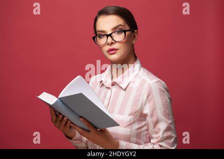 Nette Frau in einer Brille, die ein Buch auf einem roten, Studioportrait liest Stockfoto