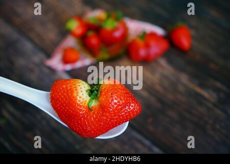 Eine einzigartige herzförmige Bio-Erdbeere auf einem weißen Löffel. Erdbeeren mit rot-weiß karierter Tischdecke auf einem rustikalen Holztisch im Hintergrund. Stockfoto