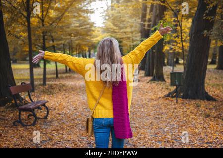 Fröhliche Frau mit gelbem Pullover, rotem Schal und Jeans. Öffentlicher Herbstpark. Schöne, sorglose junge Frau genießt den indischen Sommer. Stockfoto