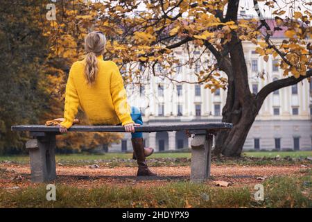 Frau, die sich in der Herbstsaison auf der Parkbank entspannt und auf Schloss schaut, Kromeriz, Tschechische Republik. Blonde Frau sitzt auf einer Bank. Stockfoto