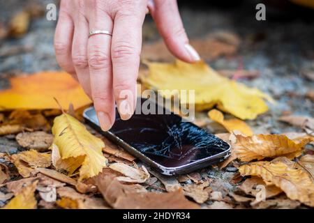 Gebrochenes Smartphone auf der Straße in gefallenen Blättern. Frau, die ein beschädigtes Mobiltelefon vom Boden pflückte. Stockfoto