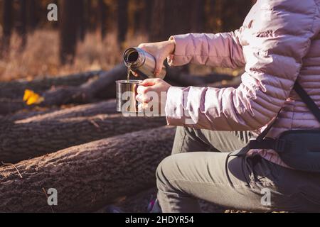 Frau gießt heißen Tee aus Thermoskannen in Metallbecher im Herbstwald. Erfrischung beim Wandern in der Natur. Stockfoto