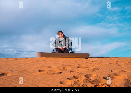 Mann mit Sandbrett sitzt auf Sand in der Wüste gegen bewölkten Himmel Stockfoto