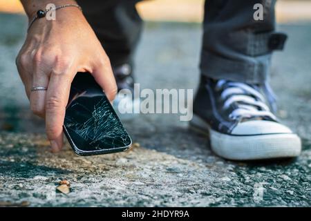 Frau nimmt zerbrochenes Smartphone vom Boden. Beschädigtes Mobiltelefon mit gesprungenem Touchscreen. Stockfoto