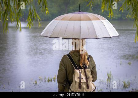 Junge Frau mit Regenschirm, die während einer Flut auf den steigenden Flussspiegel schaut. Regenzeit. Weibliche Touristin mit Rucksack im Regen. Stockfoto