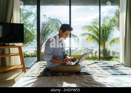 Ein junger asiatischer Geschäftsmann sitzt im Bett und arbeitet während eines Sommerurlaubs im Zimmer des Resorts in der Nähe des Meeres mit einem Laptop. Stockfoto