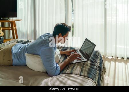 Ein junger asiatischer Geschäftsmann liegt im Bett und arbeitet während eines Sommerurlaubs im Zimmer des Resorts in der Nähe des Meeres mit einem Laptop. Stockfoto