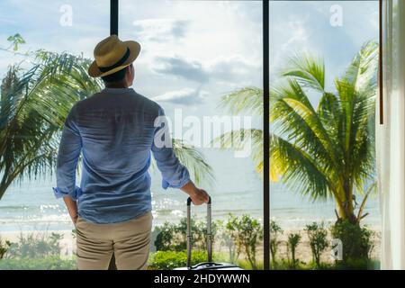 Ein asiatischer Mann mit Koffer steht neben dem Fenster seines Zimmers und blickt während seiner Sommerurlaubsreise auf den wunderschönen Meerblick. Stockfoto