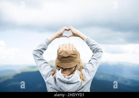 Frauen, die mit ihren Händen Herzform gegen das atemberaubende Bergpanorama der Karpaten Formen. Reisende auf dem Berggipfel genießen Luftaufnahme Hände angehoben Stockfoto