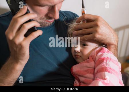 Ein fürsorglicher einsamer Vater tröstet ein krankes Mädchen, nachdem er eine hohe Temperatur mit einem Thermometer genommen hat, und ruft einen Arzt Stockfoto