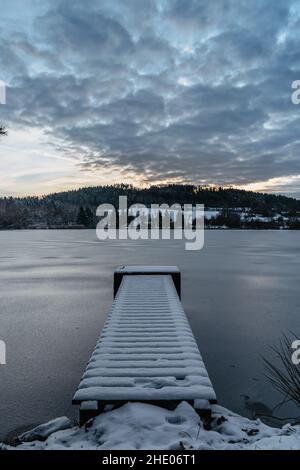 Hölzerner Pier am See mit frischem Schnee.Winterteich mit kleinem Steg bei Sonnenaufgang, Wald im Hintergrund.Frostige ruhige Landschaft. Weiße Winterlandschaft verschneit Stockfoto