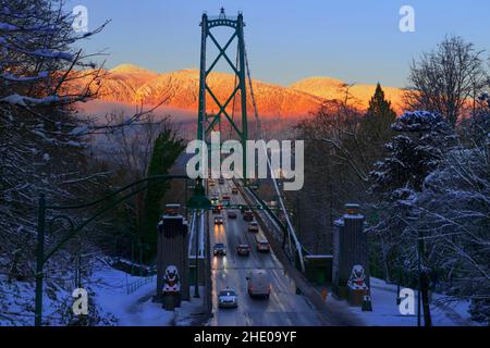 Draufsicht auf die Brücke und fahrende Autos mit Scheinwerfern, orangefarbene Berge, die von der untergehenden Sonne beleuchtet werden, blauer Himmel, schneebedeckte Bäume Stockfoto