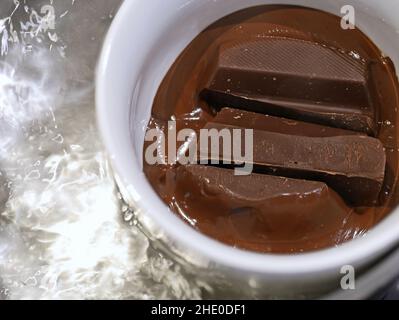 Schmelzen von dunkler Schokolade in weißer Tasse in kochendem Wassertopf, Nahaufnahme Stockfoto
