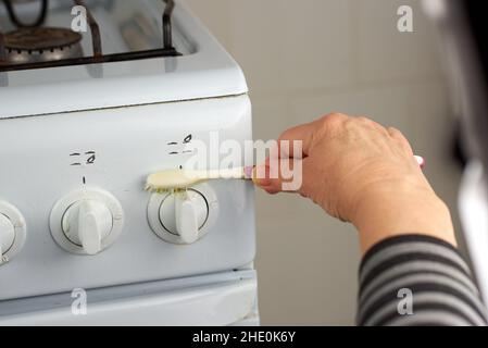 Weibliche Hand wäscht die Wasserhähne des weißen Gasofens mit einer Zahnbürste. Eine ungewöhnliche Art der Hausreinigung Stockfoto