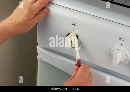 Weibliche Hände waschen die Wasserhähne des weißen Gaskochens mit einer Zahnbürste. Eine ungewöhnliche Art der Hausreinigung Stockfoto