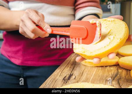 In einer Küche sieht man, wie die Hände einer jungen Frau Mayonnaise auf ein Hamburgerbrötchen legen. Stockfoto