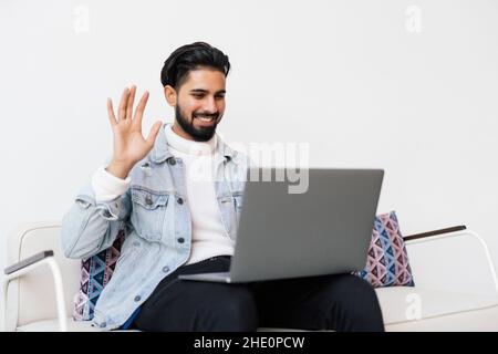 Glücklicher, tausendjähriger Geschäftsmann in der Freizeit, der Videoanruf von der Couch aus macht, die Kamera anschaut, lächelt, hallo mit der Hand winkt, an Online-Chat teilnimmt, virtu Stockfoto