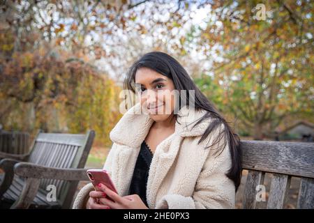 Mädchen, die auf einer Bank in einem Park sitzt, benutzt ihr Telefon und lächelt, eine attraktive indische Frau draußen während eines Herbsttages Stockfoto