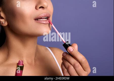 Nahaufnahme. Beschnittene Ansicht der attraktiven jungen Frau mit perfekt sauberer Haut Anwendung Lipgloss hygienischen Balsam auf dicke Lippen isoliert auf lila Hintergrund Stockfoto