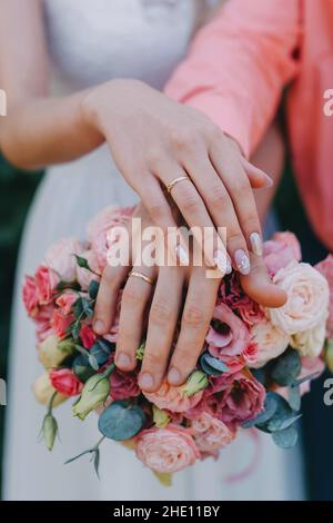 HONIGMONDE HÄNDE AUF EINEM HOCHZEITSSTRAUSS AUS BLUMEN. Hände und Ringe auf dem Brautstrauß. Braut und Bräutigam halten sich bei einer Hochzeit mit goldenen Eheringen an die Hand Stockfoto
