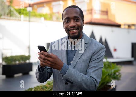 Afrikanischer Geschäftsmann lächelt, als er sein Smartphone benutzt. Glücklicher Unternehmer. Positiver Mitarbeiter mit einem inhaltlichen Gesichtsausdruck Stockfoto