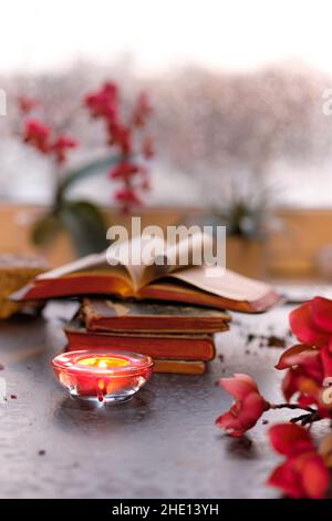 Winterzeit mit Kerze auf Stapel alter Bücher. Sonnenuntergangsfenster mit orangefarbenem Glanz, rosa und fuchsiafarbener Orchidee, Magnolienblüten. Aromatische Kerzen, Teelicht Stockfoto