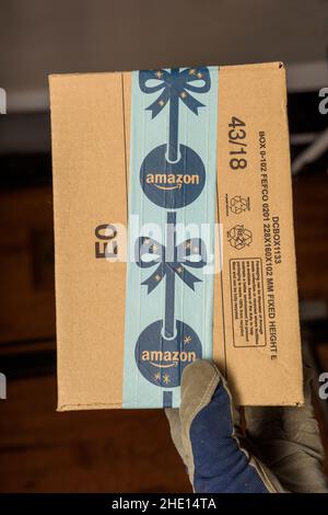 Frankfurt, Deutschland - 10. Dez 2021: Männlicher Handschuh mit Schutzhandschuh, der im Lager Amazon Karton mit weihnachtlichen Logotypien hält, während der jährlichen Wintereinkäufe in der E-Commerce-Weihnachtszeit Stockfoto