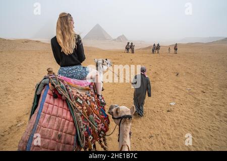 Eine alleinreisende Frau auf einem Kamelritt in der Wüste mit den Pyramiden von Gizeh, die durch starken Smog sichtbar sind. Stockfoto