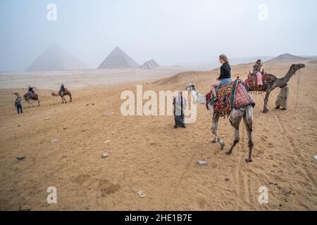 Eine alleinreisende Frau auf einem Kamelritt in der Wüste mit den Pyramiden von Gizeh, die durch starken Smog sichtbar sind. Stockfoto