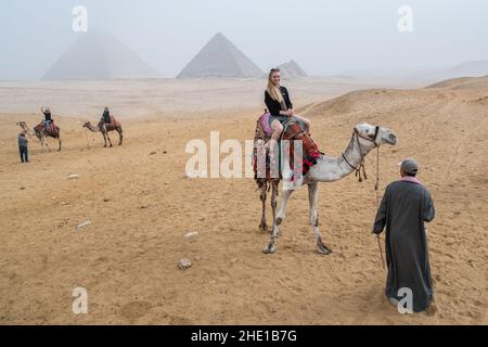 Eine alleinreisende Frau auf einem Kamelritt in der Wüste mit den Pyramiden von Gizeh, die durch starken Smog sichtbar sind. Stockfoto