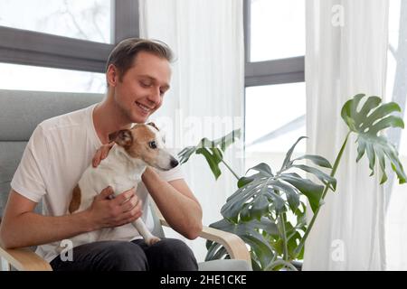 Sanfter positiver männlicher Besitzer, der im Sessel sitzt und den entzückenden Jack Russell Terrier neben dem Fenster und dem Blumentopf mit Monstera streicheln kann Stockfoto