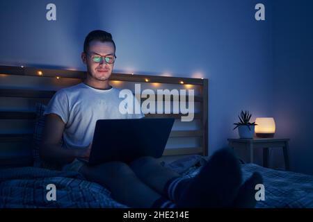 Mann mit Brille sitzt im Schlafzimmer auf dem Bett und benutzt einen Laptop. Das Gesicht wird vom Computerdisplay beleuchtet. Stockfoto