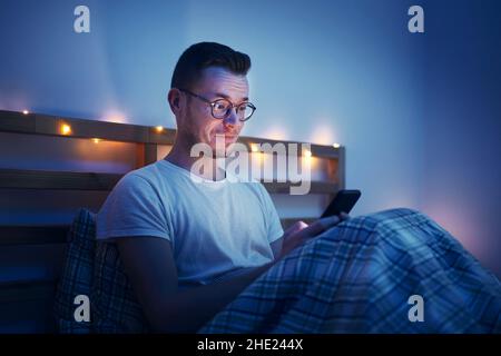Mann mit Brille sitzt unter der Bettdecke auf dem Bett im Schlafzimmer und benutzt Telefon. Überraschendes Gesicht vom Computerdisplay beleuchtet. Stockfoto