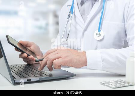 Der Arzt tippt auf dem Laptop, während er am Glastisch im Krankenhausbüro sitzt. Nahaufnahme eines unbekannten männlichen Arztes, der im Krankenhaus und am Tisch sitzt Stockfoto