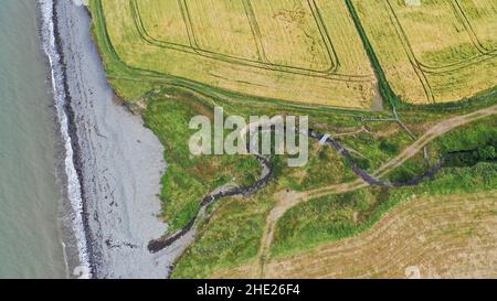 Luftbild eines gewundenen Baches, der zum Meer fließt, können Sie Felder, Strand und Meer sehen. Der Küstenweg führt über den Bach über eine große Schieferplatte. Stockfoto