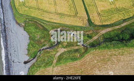 Luftbild eines gewundenen Baches, der zum Meer fließt, können Sie Felder, Strand und Meer sehen. Der Küstenweg führt über den Bach über eine große Schieferplatte. Stockfoto