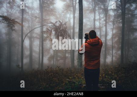 Junger bärtiger Wanderer in orangefarbener Jacke, der mit Nebel und Nebel Fotos im Wald gemacht hat. Rückansicht des Fotografen im Feld, der die Aussicht genießt. Stockfoto