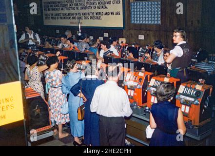 Die Kunden an den Pokerautomaten im Harold's Club, einem Casino in der North Virginia Street, Reno, Nevada, USA, wurden Ende 1950s angesehen. Eine Glücksspielmaschine, wie sie hier gespielt wird, wird oft als ‘einarmiger Bandit’, ‘Spielautomat’, ‘Fruchtautomat’ oder ‘Pokey’ bezeichnet und Menschen ‘spielen’ die Früchte, Spielautomaten oder Pokies. Ein Plakat an der Wand zeigt an, dass, selbst wenn Spieler Geld verlieren, einige an einen guten Zweck gehen werden – Stipendien an der Universität von Nevada. Dieses Bild stammt von einem alten amerikanischen Amateur Kodak Farbtransparenz - ein Vintage 1950s Foto. Stockfoto