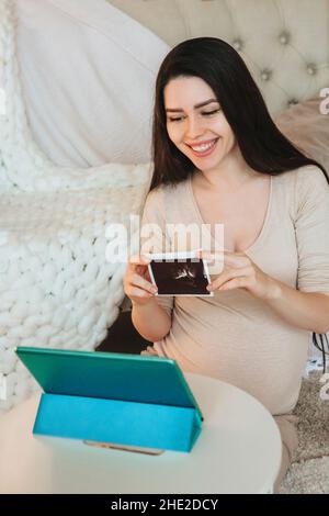 Junge glückliche Schwangere Frau mit Baby-Ultraschall in der Hand, die online auf einem digitalen Tablet spricht, Videoanruf mit der Familie führt, lächelnde Frau, die Sonogras zeigt Stockfoto