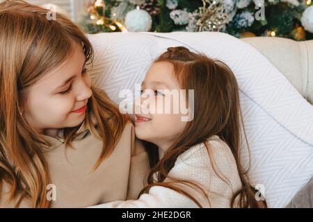 Gemütliches häusliches Porträt zweier lächelnder kleiner Mädchen-Schwestern, die sich umarmen, während sie zu Hause ihre Freizeit verbringen und auf dem Bett in der Nähe von d sitzen Stockfoto