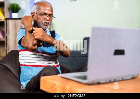 indischer älterer Mann massiert die Hand mit dem Holzrollermassager, indem er sich von zu Hause aus online Medikationsvideos auf dem Laptop ansieht - Konzept der Physiotherapie Stockfoto