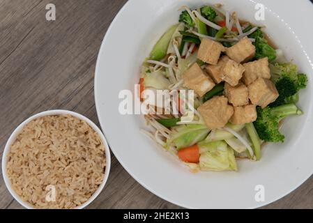 Draufsicht auf einen Haufen gemischter Vegetabel mit Tofu-Würfeln auf der Oberseite, serviert mit einer Schüssel braunem Reis. Stockfoto